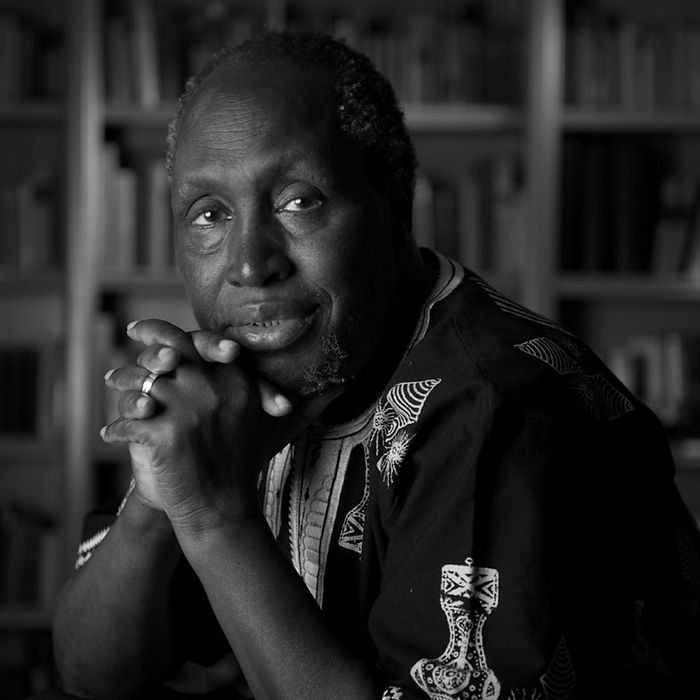 Portrait en noir et blanc d'un veil homme noir assis dans une bibliothèque en tenue africaine, les mains croisées sous le menton, un sourire en coin
