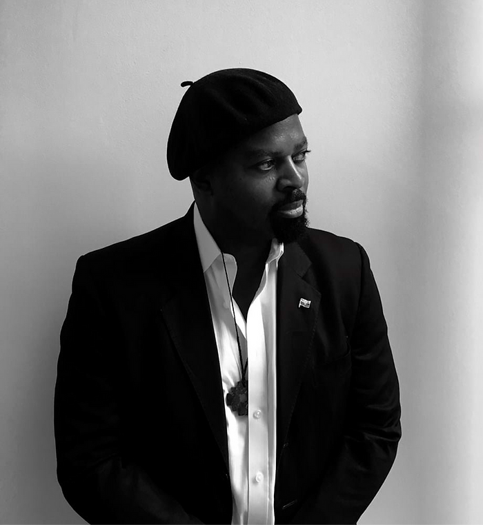 Portrait en noir et blanc d'un homme noir debout, béret sur la tête, les bras croisés